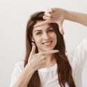 Creative girl imaging how cute her new dress will look on model. Portrait of friendly charming designer finger framing and looking through at camera with broad smile, standing over gray wall.