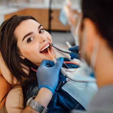 Young female patient visiting dentist office. Beautiful woman with healthy straight white teeth sitting at dental chair with open mouth during oral checkup while doctor working at teeth.