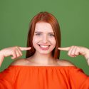 Young red hair woman in casual orange blouse on green background point fingers on white teeth perfect smile